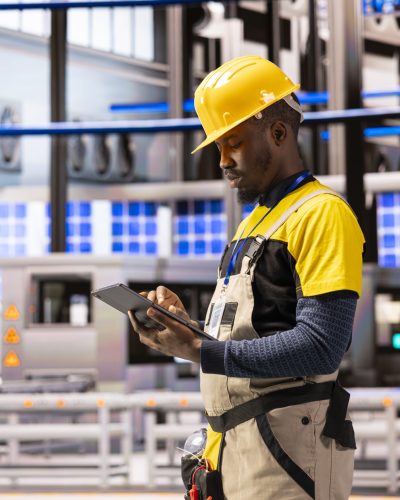 Male engineer analyzing industry 4.0 system performance in smart manufacturing plant. Black man monitoring machinery and robotic arms, ensuring optimal production and industrial automation efficiency.